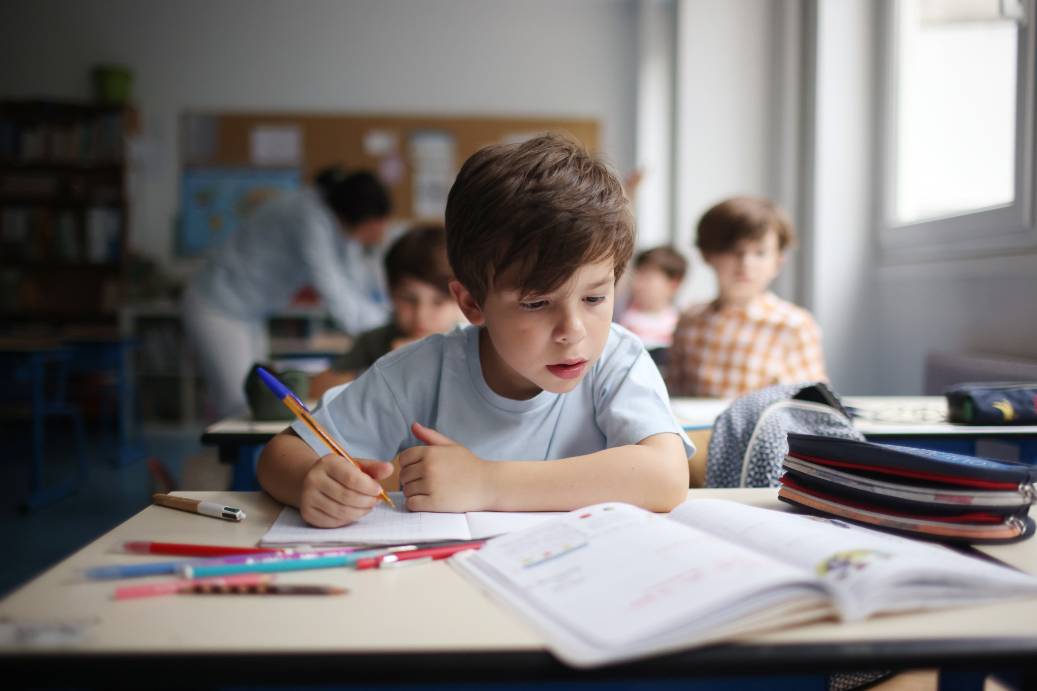 Ein Junge konzentriert sich auf seine Schulaufgaben, umgeben von Klassenkameraden und Unterrichtsmaterialien.