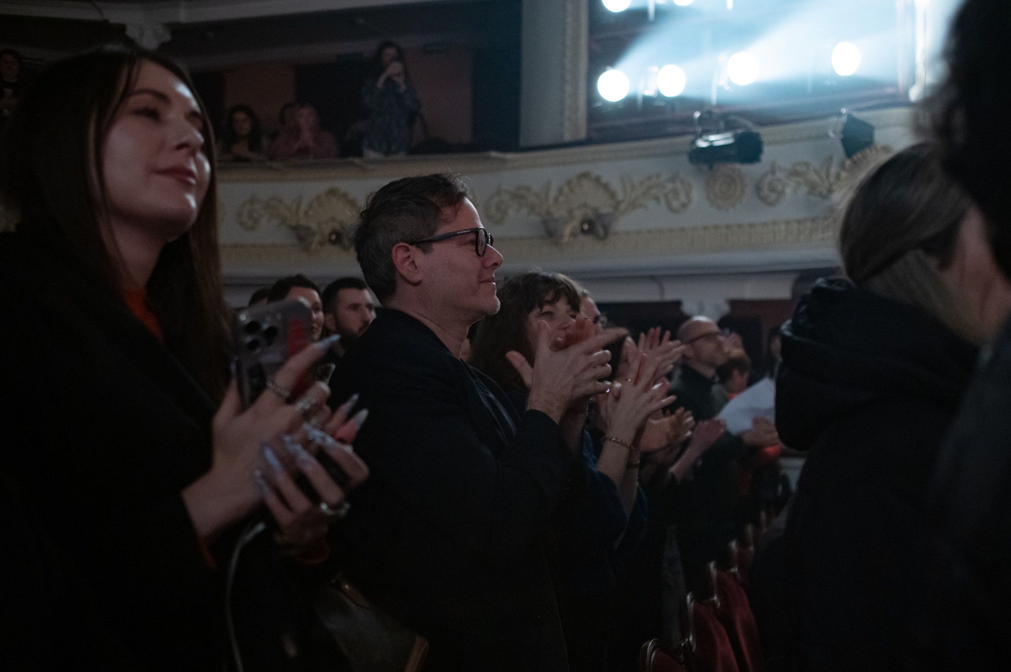Menschen in einem Theater applaudieren während einer Vorstellung, im Hintergrund leuchten Scheinwerfer.